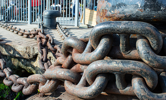 Bollard and chain close-up. They have a black color and some parts are rusty. It is used to fix the ship to the port. Photographed on a sunny day.
