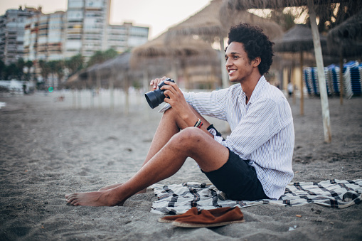 Handsome, young photographer with afro hairstyle sitting on the beach and holding digital camera.