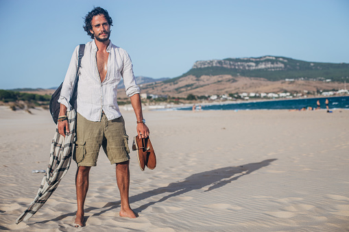 Handsome, modern man with bag standing on the beach and holding shoes.