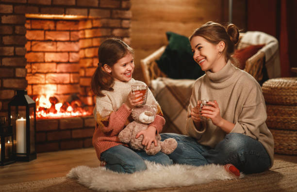 family mother and child reading book and drink tea on winter evening by fireplace family mother and child daughter reading book and drink tea on winter evening by fireplace young hot stock pictures, royalty-free photos & images