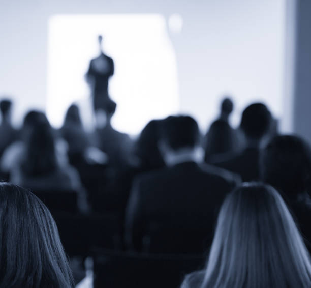 istock Conference photo audience and speaker giving speech. Seminar presenter on a panel during forum. Corporate manager in sales executive training discussion on stage. Investor pitch presentation. 1203554968