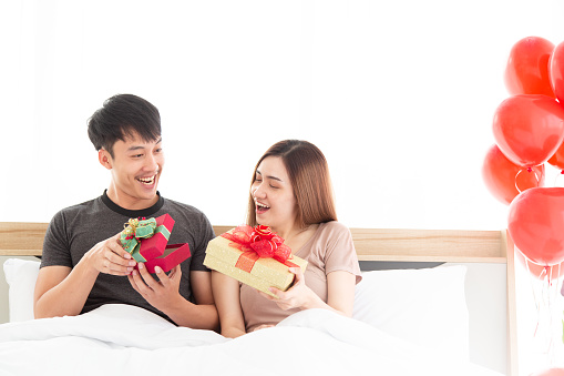 Happy and excited young adult couple opened present gifts boxes, spending weekend at home, sitting on bed with red balloons. Valentine's Day. Wedding or Birthday Anniversary.