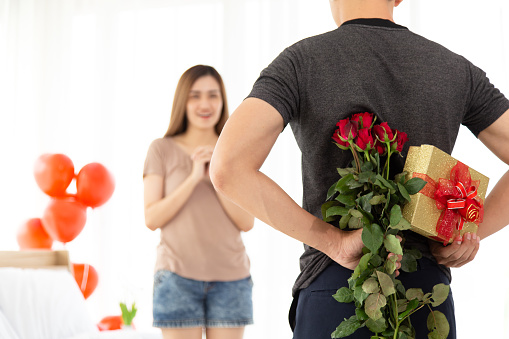 Young man's hands hiding holding chic bouquet of red roses and gift with red ribbon behind back, happy woman is on blurred background. Celebrate anniversary, love & relationship in valentine's day.