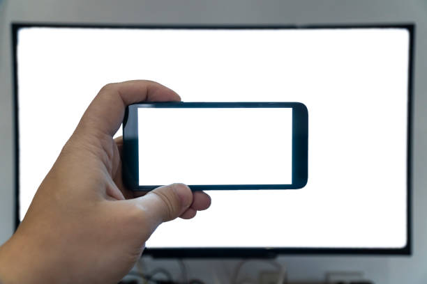 Living through the screen concept. Man holding a smartphone in front of a ultra hd tv. Both with blank screens.