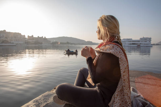Mature woman sits on a ghat at sunrise with a hot beverage, looks out across lake Lake Pichola, Udaipur lake palace stock pictures, royalty-free photos & images