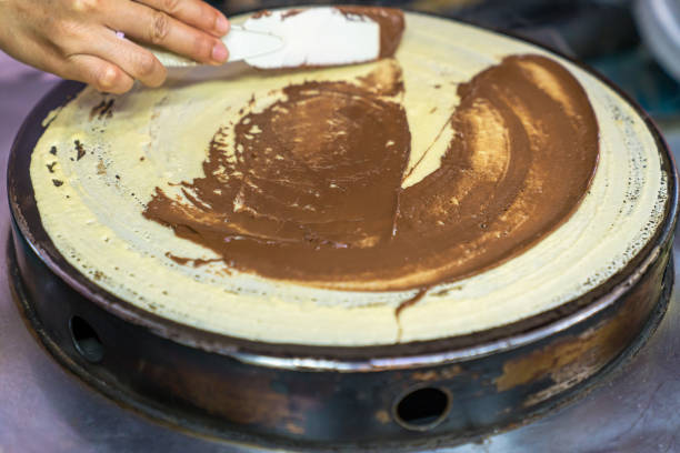 istock Thai pancakes preparation: the cook gently smearing and spreading chocolate paste on the pancakes being fried on the pancake maker. street food, close-up 1203573555