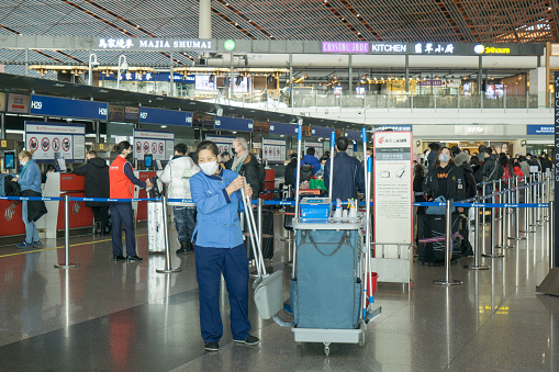 Beijing, China, January 31, 2010:  Beijing Capital International Airport during the outbreak of Corona virus. The virus has rapidly spread and became a global health emergency. Passengers wearing masks wait in line at the check in counters of Air China. In the foreground is a janitor cleaning the floor.