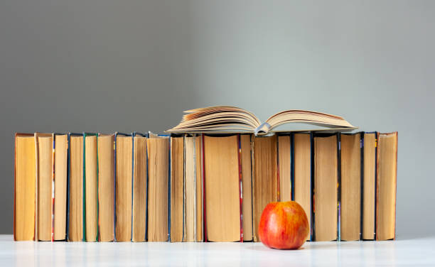 istock Open hardback book, diary, fanned pages on stack of books on white table with red apple. Books stacking. Back to school concept. Copy Space. Education learning background 1203580705