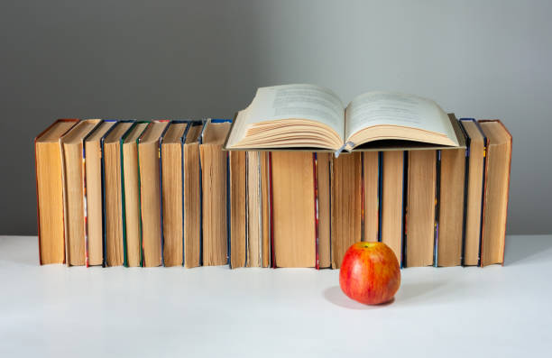 istock Open hardback book, diary, fanned pages on stack of books on white table with red apple. Books stacking. Back to school concept. Copy Space. Education learning background 1203580734