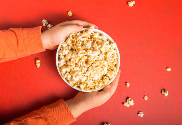 istock Popcorn viewed from above on red background. Child eating popcorn. Human hand.  Cinema snack concept. The food for watching a movie and entertainment. Copy space for text, flat lay. 1203602064
