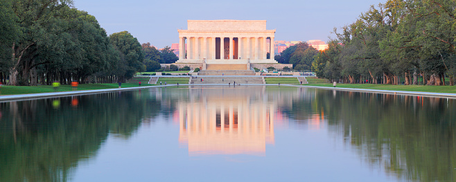 The picture shows the Lincoln Memorial, an iconic neoclassical monument located in Washington D.C., USA. It is a bright, clear day with blue skies. The memorial has a large number of white stone columns supporting the structure. In front of the memorial, there are steps leading up to the entrance where we can see visitors gathered and moving about. The structure exudes a sense of grandeur and historical significance, and it appears to be a popular tourist location. There are a few people in the foreground at the bottom of the steps, suggesting the photo was taken from a considerable distance to capture the full facade of the building.