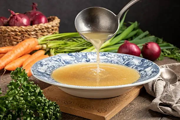 Pouring chicken bone broth from a ladle into a vintage plate, with fresh vegetables in the background