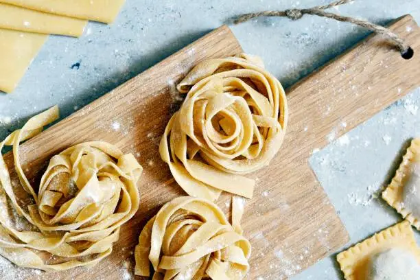 Homemade italian pasta, ravioli, fettuccine, tagliatelle on a wooden board and on a blue background. The cooking process, raw pasta. Tasty raw ravioli with ricotta and spinach,with flour on background