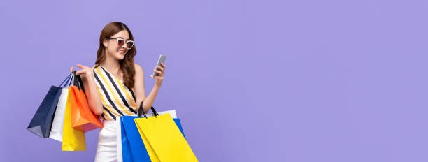 Beautiful Asian woman shopping online with mobile phone on banner background Trendy beautiful young Asian woman carrying colorful bags shopping online with mobile phone isolated on purple banner background shopping bag photos stock pictures, royalty-free photos & images