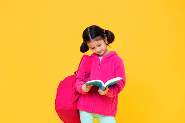 Portrait of cute pretty girl with pigtail hair in pink jacket and backpack reading book in yellow isolated studio background