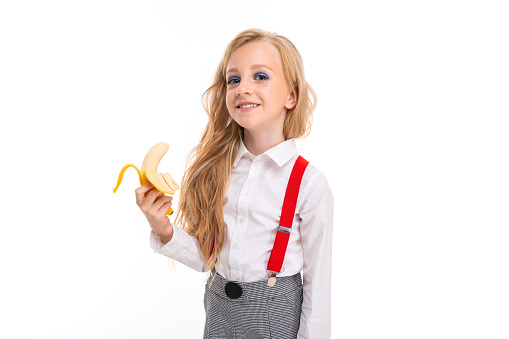 Little girl with long blonde hair in white shirt, red pull-ups, pants in cage, red socks and bolts with bright makeup with banana