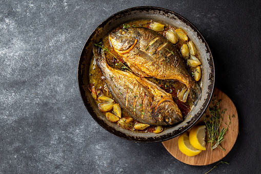 Baked sea bream or dorada with onion and herbs in pan on dark background.