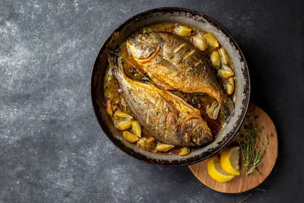 Photo of Baked sea bream or dorada with onion and herbs in pan on dark background