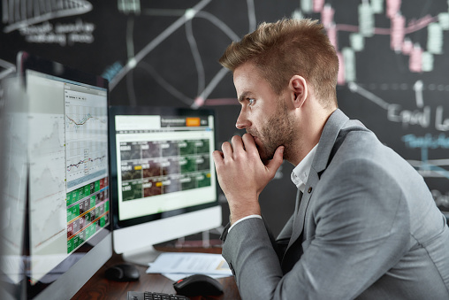 Portrait of successful trader looking focused while sitting in front of monitors in the office. Blackboard full of charts and data analyses in background. Stock trading, business concept. Side view