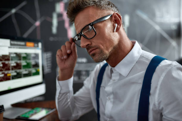 cuide do seu futuro. feche de acordo com o empresário elegante, comerciante sentado na frente do monitor de computador enquanto trabalha usando fones de ouvido sem fio. quadro negro cheio de gráficos e análises de dados em segundo plano. - stock certificate audio - fotografias e filmes do acervo