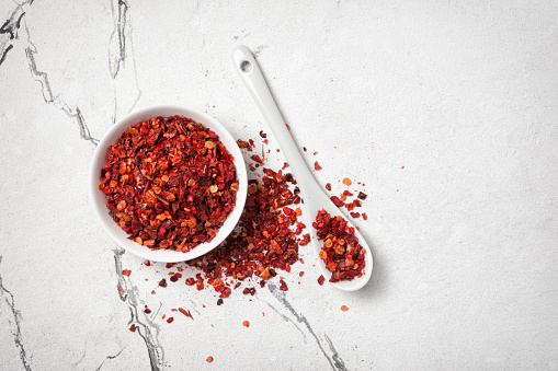 Top view of dried hot red pepper flakes in bowl for spicy food on white marble background with copy space