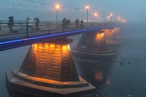 Uzhgorod, Ukraine - January 18, 2020: Fuzzy silhouettes of people passing through a pedestrian bridge in dense evening fog. Mystical background.