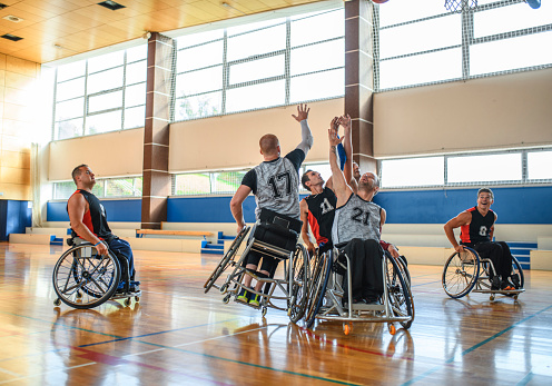 Male wheelchair basketball players aggressively reaching for control of rebound during practice game.