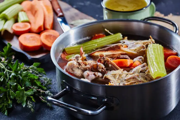 cooked chicken stock with vegetables and aromatic herbs in a stockpot and in a mug at the background, ingredients on a stone kitchen worktop, horizontal view, close-up