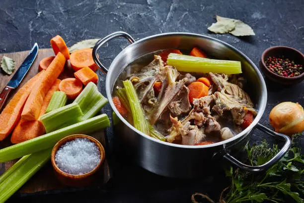 cooked chicken stock with vegetables and aromatic herbs in a stockpot, ingredients on a kitchen worktop, close-up