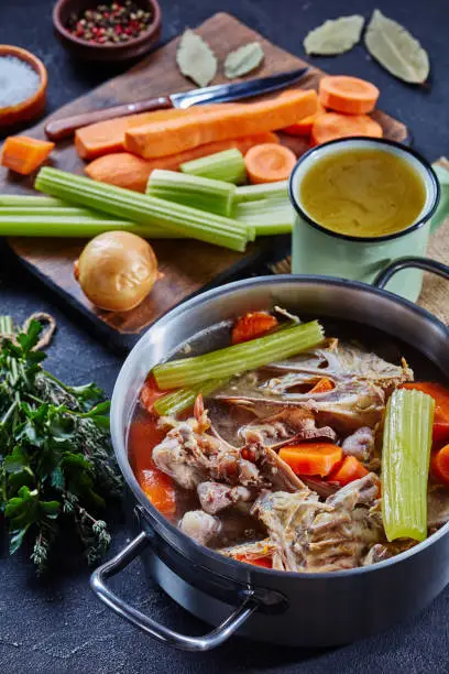 cooked chicken stock with vegetables and aromatic herbs in a stockpot and in a mug at the background, ingredients on a stone kitchen worktop, vertical view, close-up