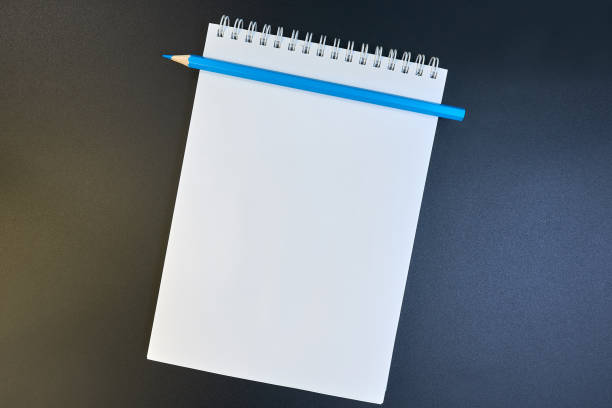 istock Blue pencil on a white sheet of notepad paper on a dark gradient from brown to blue background. Blank paper sheet and pencil for mock up. copy space 1203692568