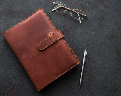 Brown leather notebook, pen and glasses on black background