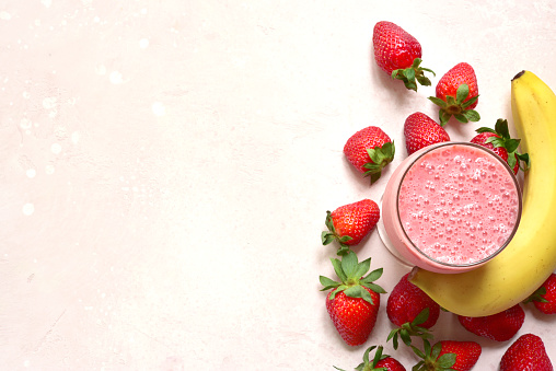 Delicious strawberry banana smoothie in a glass with ingredients for making on a light pink slate, stone or concrete background. Top view with copy space.