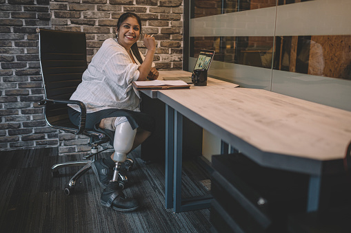 an asian indian female white collar worker with prosthetic leg sitting in front of her work station in office smiling and working with her laptop and coffee cup in the morning