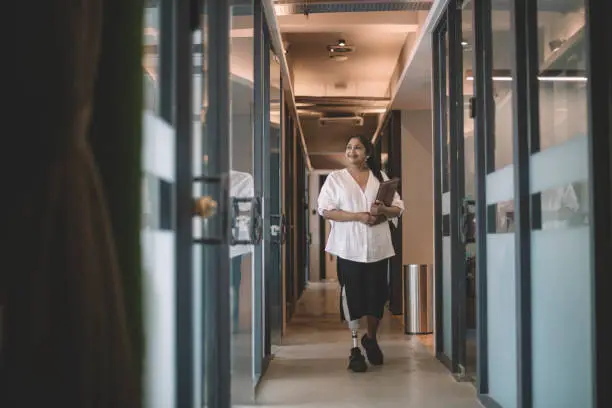 an asian indian female handicapped white collar worker with prosthetic limb  holding her digital tablet and walking at the office corridor smiling
