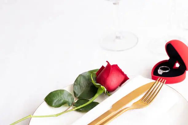 Red rose on white plate with gold cutlery and red box with engagement ring . Saint valentines day celebration or propose romantic dinner concept.Copy space
