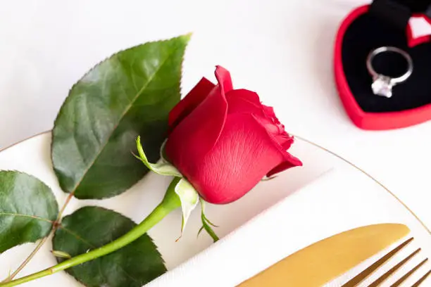 Red rose on white plate with gold cutlery and red box with engagement ring . Saint valentines day celebration or propose romantic dinner concept.