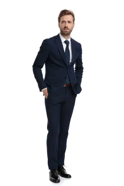 concerned young businessman in navy blue suit holding hands in pockets, looking to side and walking isolated on white background, full body