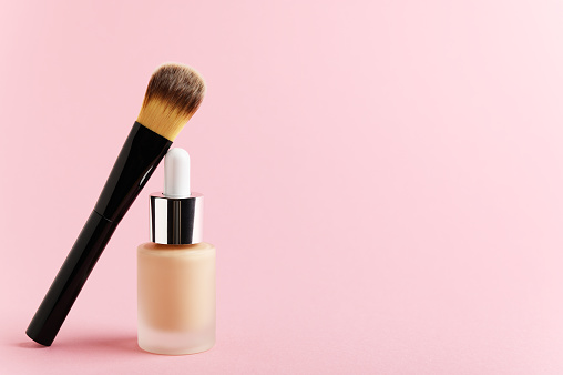Liquid foundation cream unbranded bottle with makeup brush. Facial correction, liquid concealer, tone, bb, cc cream skincare product on pink background. Feminine cosmetics accessory with copy space.