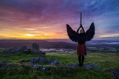 A lone Black Winged Warrior Angel holding a sword overlooking beautiful British landscape at dawn