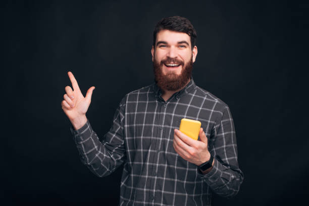 istock Handsome bearded guy is pointing aside and holding his phone in the other hand. 1203730627