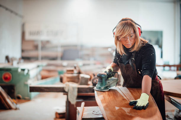 Woodworker using a hand sander to sand down a wooden surface Girl carpenter using an orbit sander to sand down a wooden panel on a work bench in a workshop. blue collar worker photos stock pictures, royalty-free photos & images