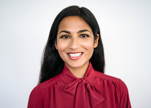Front view close-up of young Indian businesswoman with long black hair wearing maroon blouse with tied bow and smiling at camera against white background.