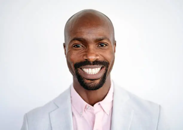 Front view close-up of African businessman with goatee wearing pale gray suit jacket over pink button down shirt and smiling at camera against white background.