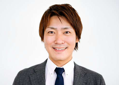 Front view close-up of young Japanese business executive wearing gray suit jacket over shirt and tie and smiling at camera against white background.