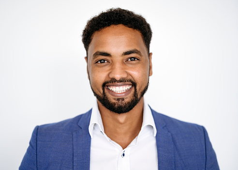 Front view close-up of casual bearded mid adult African businessman wearing blue blazer over open collar shirt and smiling at camera against white background.