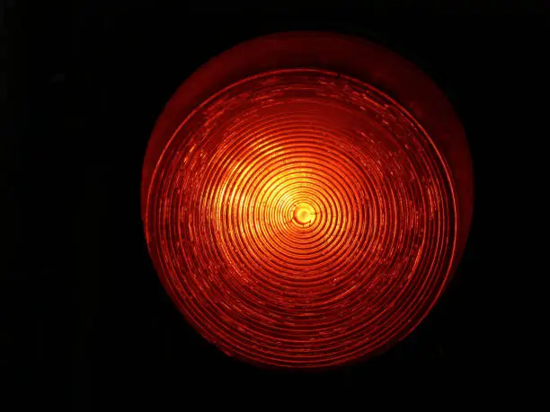 isolated red traffic light closeup with yellow undertone and black background. concentric circles on bright glass lens. transportation signs and symbols concept. abstract view.