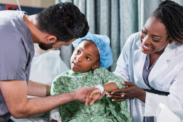 Doctor, nurse helping girl sitting on hospital bed A 6 year old African-American girl sitting on a hospital gurney, with a doctor and nurse tending to her. She is wearing a hospital gown and surgical cap. A nurse in scrubs, a young Middle Eastern man in his 20s, is standing on one side of her, and a female doctor, a mature African-American woman in her 40s, is standing on the other side. girl patient stock pictures, royalty-free photos & images