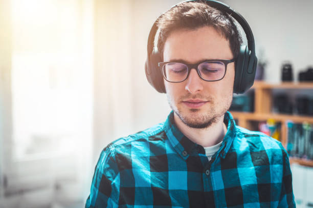 słuchanie muzyki w domu: młody kaukaski mężczyzna słucha muzyki ze słuchawkami. niebieska koszula w kratę. - muzykoterapia zdjęcia i obrazy z banku zdjęć