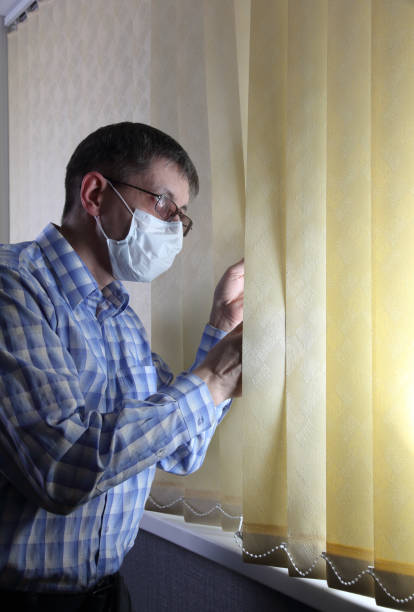 man in a medical face mask looks out the window through the blinds - russian influenza epidemic virus flu virus imagens e fotografias de stock